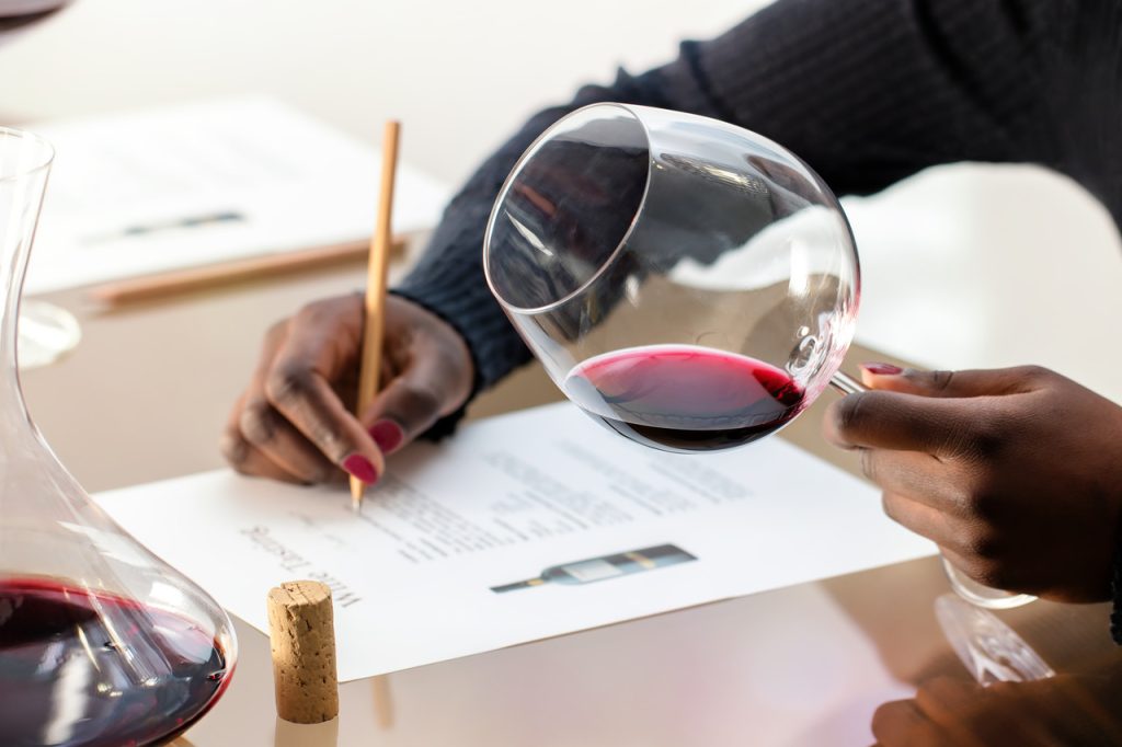 Hand of a person holding a glass of red wine while with the other hand taking notes on a sheet of paper